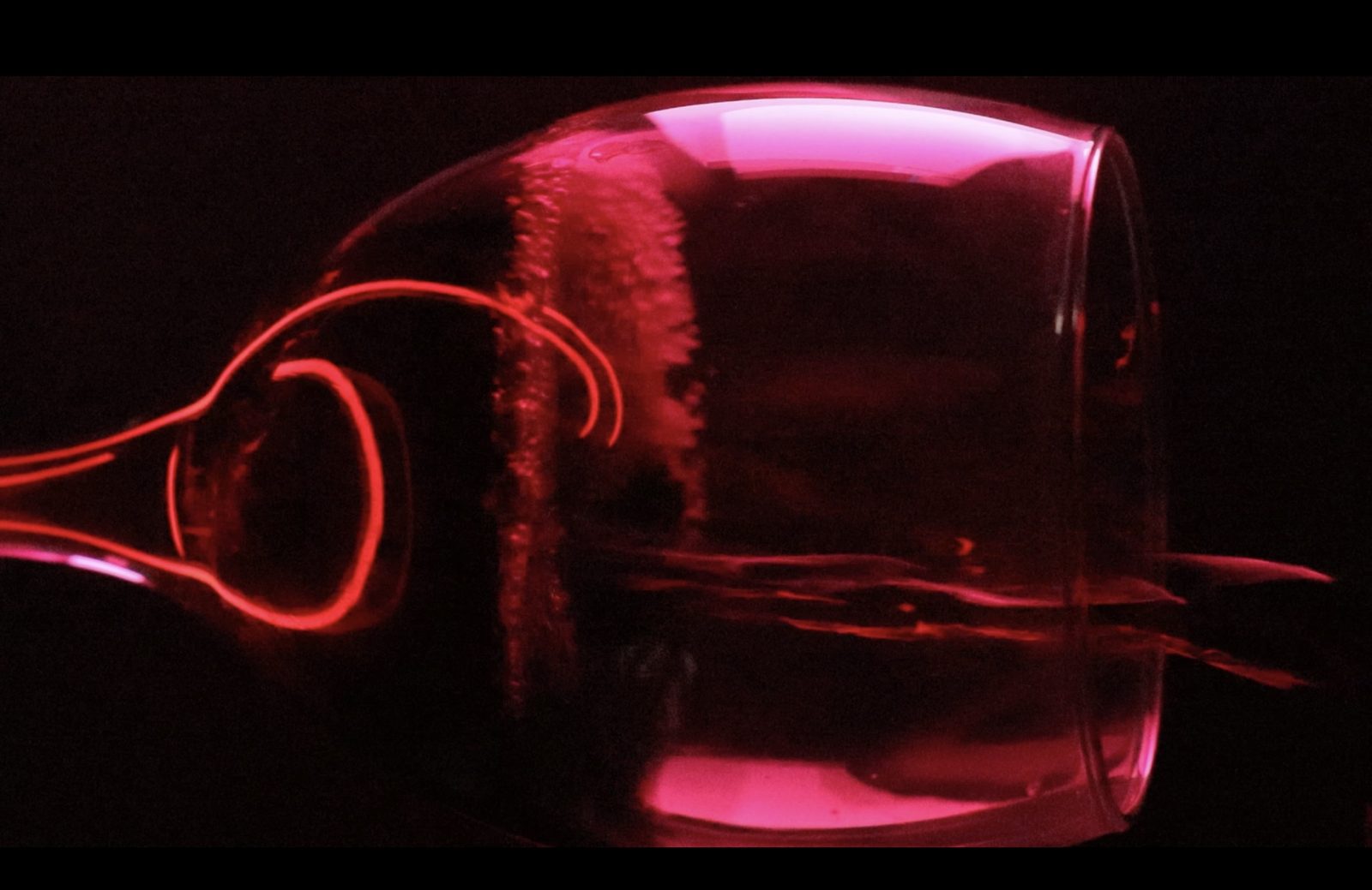 A wine glass lying on its side, lit pink and red against black.